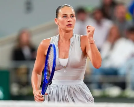 Marta Kostyuk in white outfit holds a blue racket and raises her fist in celebration