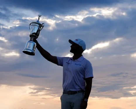 Wyndham Clark holds a trophy aloft against a dramatic cloudy sky at dusk.