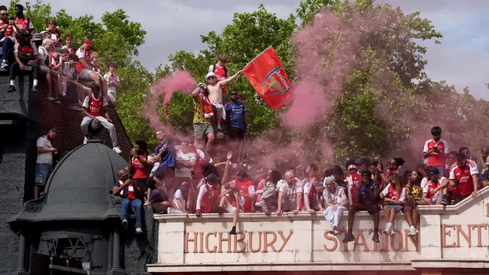 A large group of Arsenal football fans celebrate whilst gathered on the roof and structure of Highbury Station. Several supporters wear the club's red and white shirts, one fan waves a large Arsenal flag, and another holds a flare that releases thick plumes of pinkish-red smoke into the air.